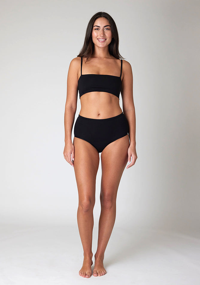 Front image of a five-foot-ten, size 10 model wearing Ugly Pants' black Overnight period pants in moderate to heavy absorbency, paired with a black crop top, this image does not feature the self tie bow, but a straight neckline.
