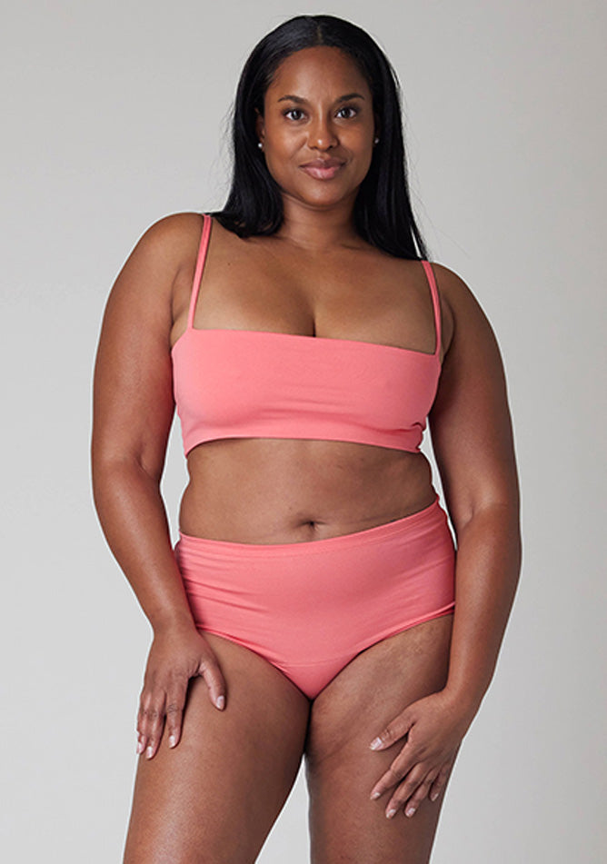 Close up image of a five-foot-five, size 10 model wearing Ugly Pants' Coral Overnight period pants in moderate to heavy absorbency, paired with a coral crop top, this image does not feature the self tie bow, but a straight neckline.