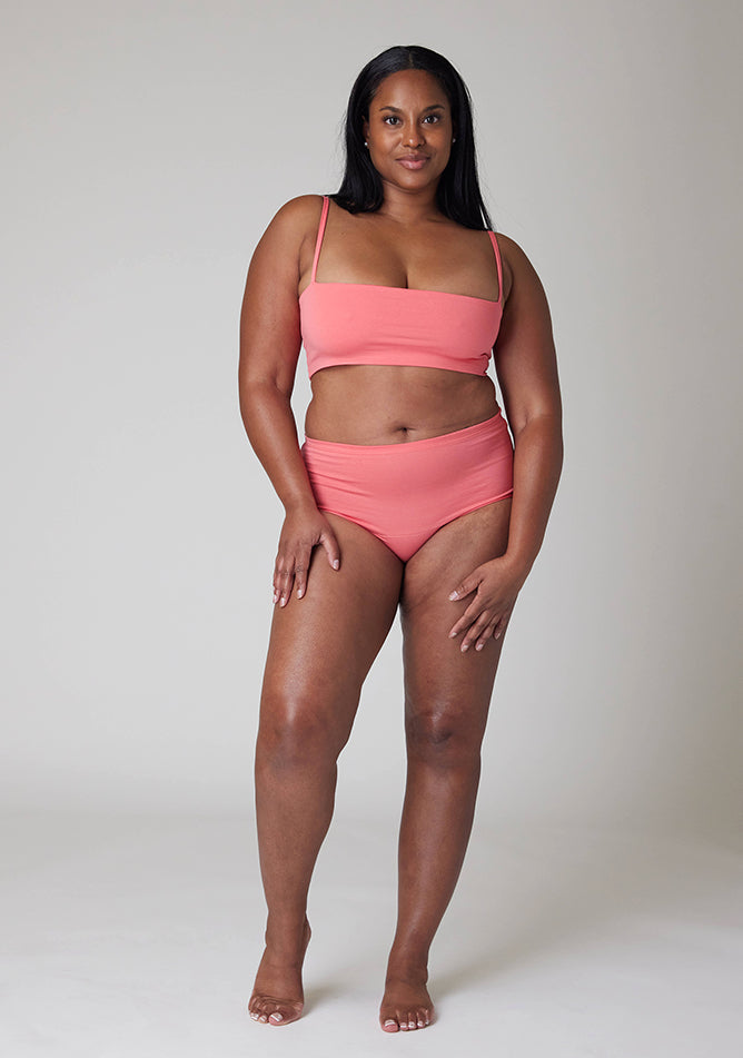 Front image of a five-foot-five, size 10 model wearing Ugly Pants' Coral Overnight period pants in moderate to heavy absorbency, paired with a coral crop top, this image does not feature the self tie bow, but a straight neckline.