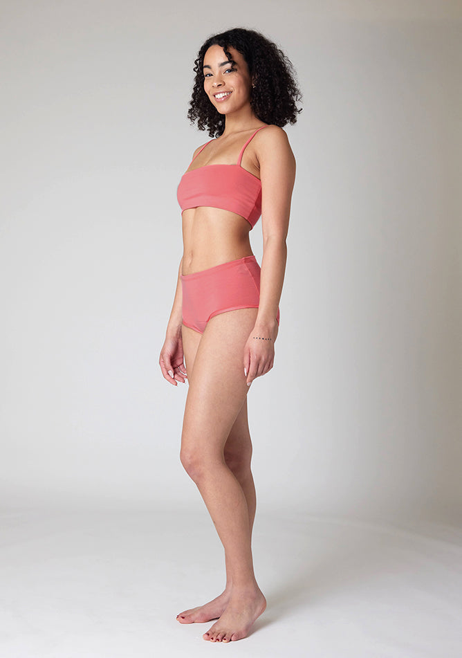 Front quarter image of a five-foot-ten, size 10 model wearing Ugly Pants' Coral Overnight period pants in moderate to heavy absorbency, paired with a coral crop top, this image does not feature the self tie bow, but a straight neckline.