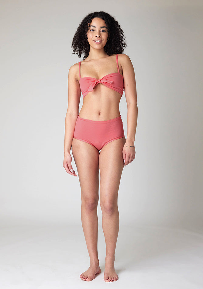 Front tied image of a five-foot-ten, size 10 model wearing Ugly Pants' Coral Overnight period pants in moderate to heavy absorbency, paired with a coral crop top, this image does not feature the self tie bow.