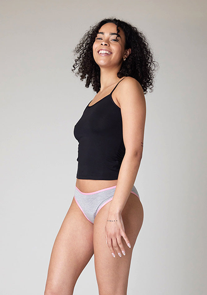 Close-up front quarter image of a five-foot-ten, size 10 model wearing Ugly Pants' Grey Bikini Brief period Pants with light pink elastic in moderate to heavy absorbency, paired with a black cami top