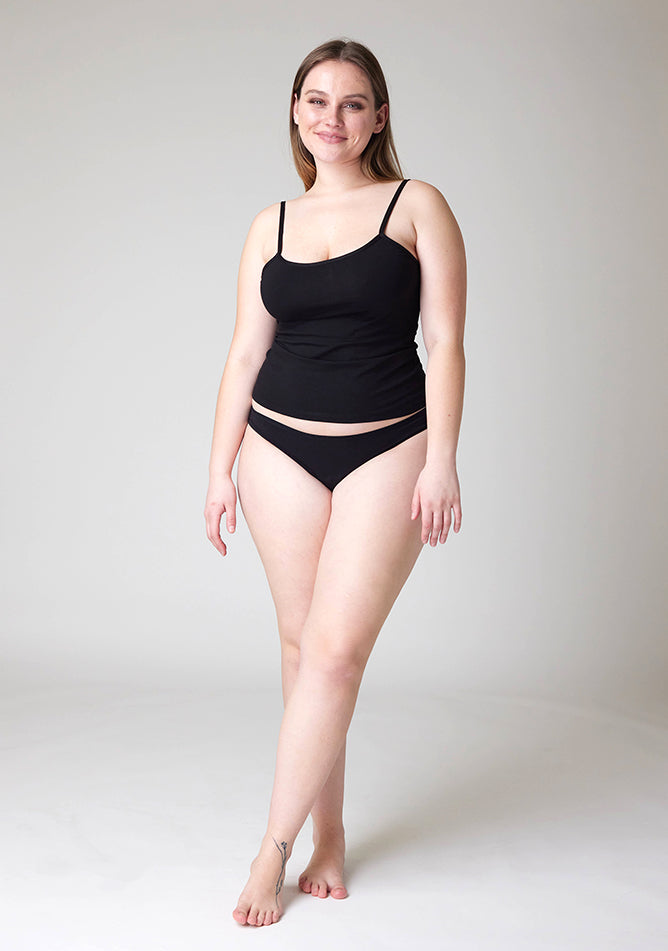 Front image of a five-foot-five, size 14 model wearing Ugly Pants' Black Bikini Brief Period Pants in moderate  to heavy absorbency, paired with a black cami top