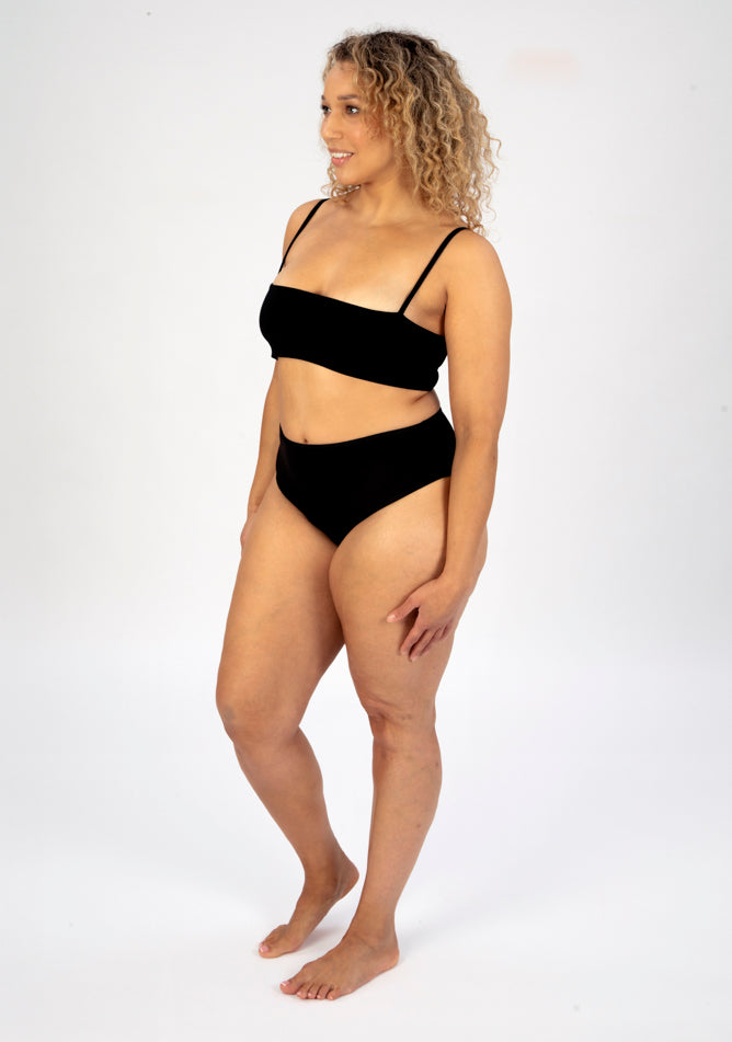 Front quarter image of a five foot seven, size 16 model wearing Ugly Pants' black high waisted period pants in light to moderate absorbency, paired with a black crop top.