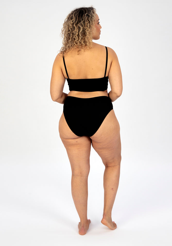 Back image of a five foot seven, size 16 model wearing Ugly Pants' black high waisted period pants in light to moderate absorbency, paired with a black crop top.