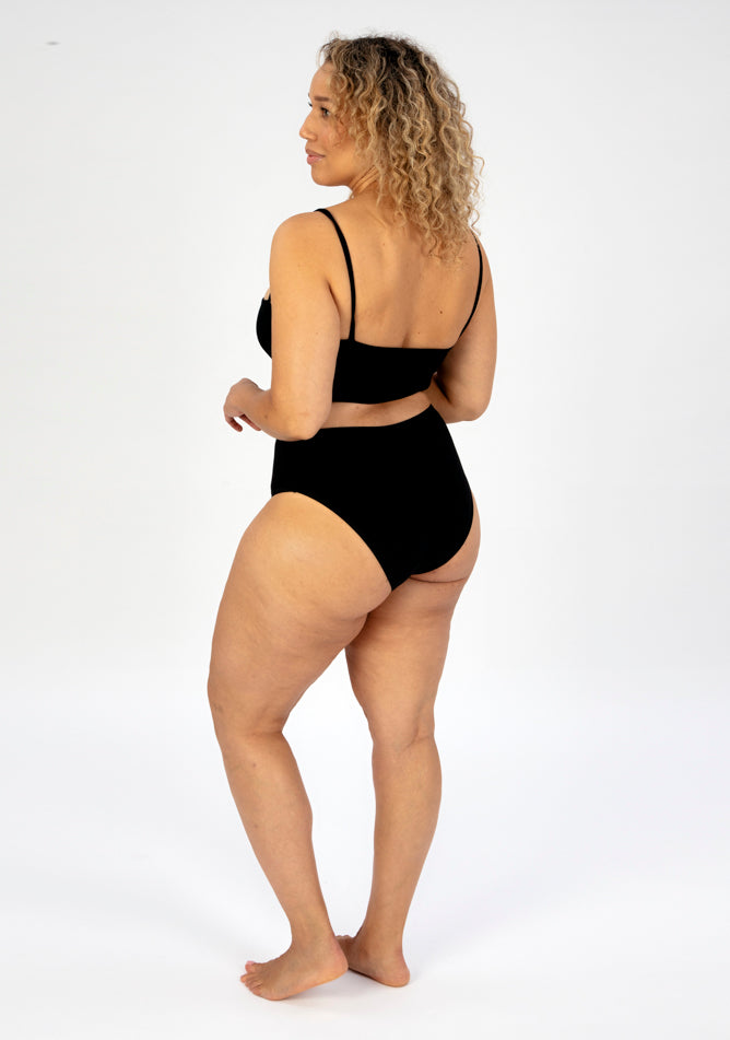 Back quarter image of a five foot seven, size 16 model wearing Ugly Pants' black high waisted period pants in light to moderate absorbency, paired with a black crop top.