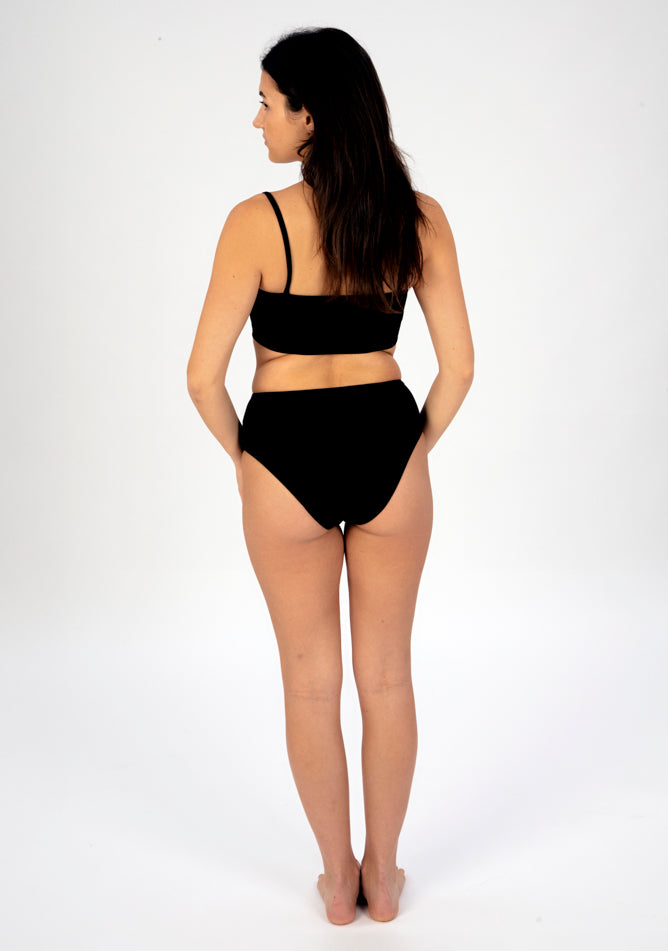 Back image of a five foot six, size 8 model wearing Ugly Pants' black high waisted period pants in light to moderate absorbency, paired with a black crop top.