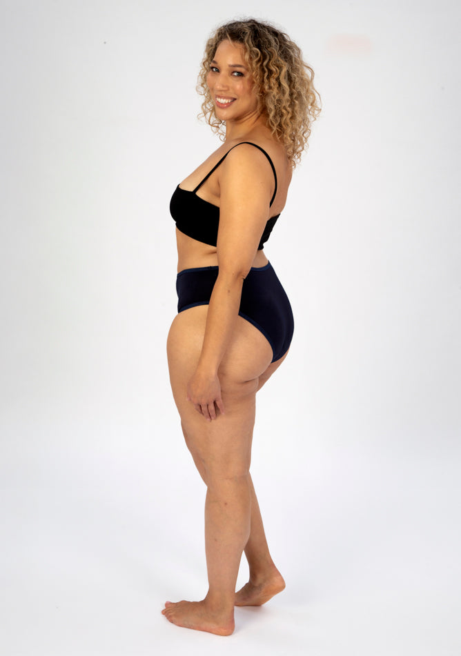 Back quarter image of a five foot seven, size 16 model wearing Ugly Pants' navy high waisted period pants in light to moderate absorbency, paired with a black crop top.