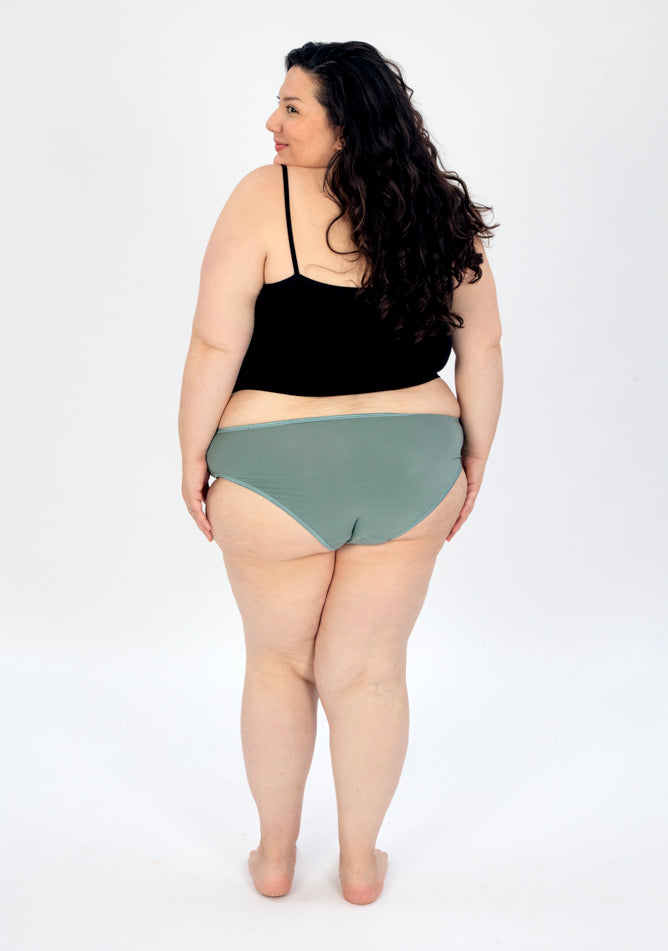 Back image of a five foot seven, size 22 model wearing Ugly Pants' sage high waisted period pants in moderate to heavy absorbency, paired with a black cami top.