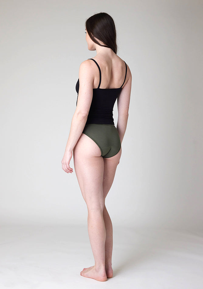 Back quarter image of a five-foot-nine, size 8 model wearing Ugly Pants' Olive Bikini Brief period Pants in light to moderate absorbency, paired with a black cami top