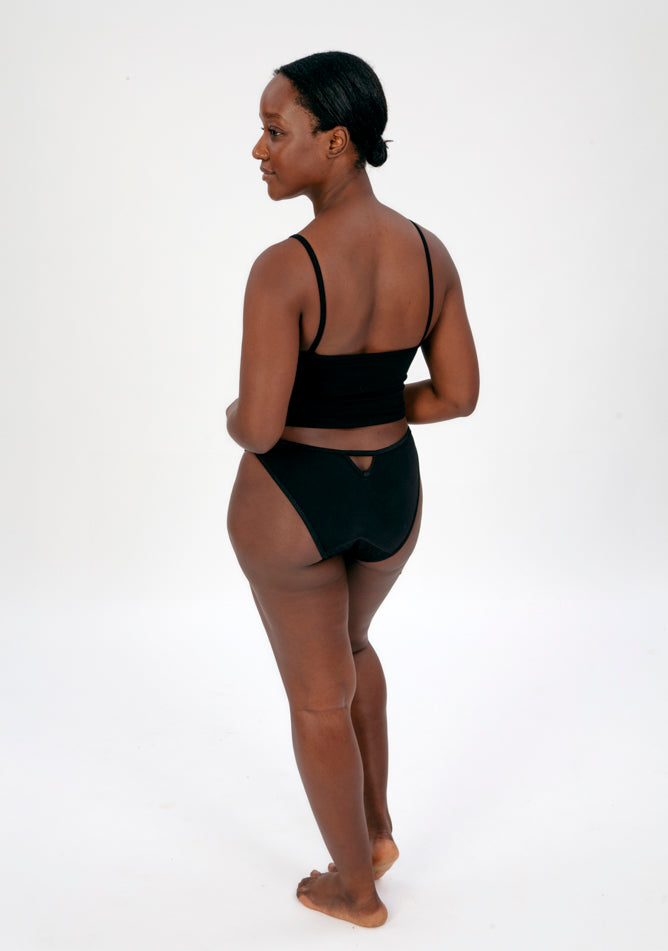 Back quarter image of a five foot four, size 10 model wearing Ugly Pants' Black Lace Bikini Brief Period Pant in a light to moderate absorbency, paired with a black crop top.
