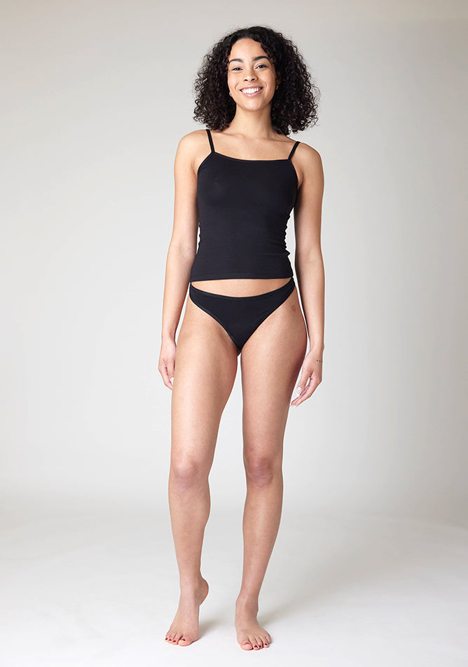 Front image of a five-foot-ten, size 10 model wearing Ugly Pants' Black Thong period pants in a light absorbency, paired with a black cami top.
