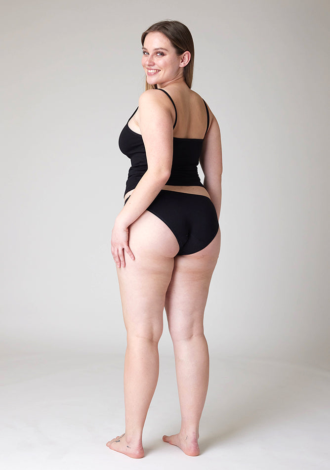 Quarter back image of a five foot five, size 14 model wearing Ugly Pants' Black Bikini Brief period pants in light to moderate absorbency, paired with a black cami top.