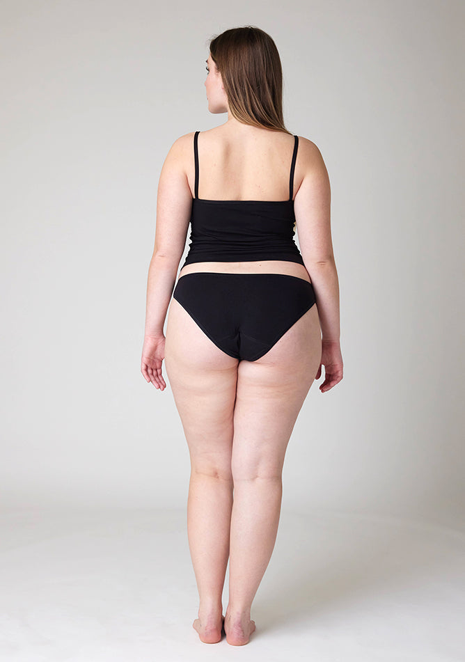 Back image of a five foot five, size 14 model wearing Ugly Pants' Black Bikini Brief period Pants in moderate  to heavy absorbency, paired with a black cami top