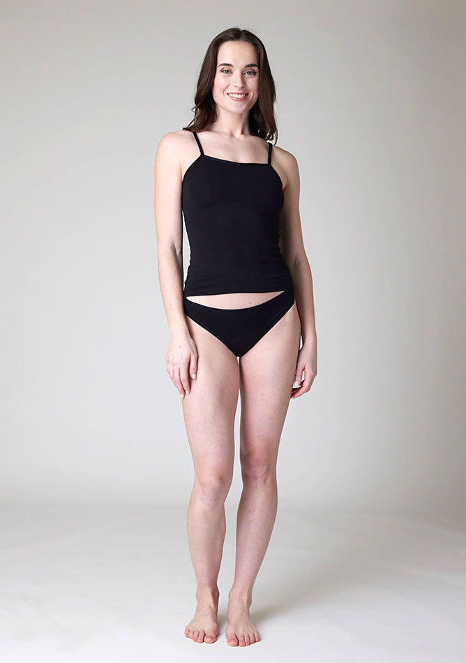 Front image of a five foot nine, size 8 model wearing Ugly Pants' Black Bikini Brief period Pants in moderate  to heavy absorbency, paired with a black cami top