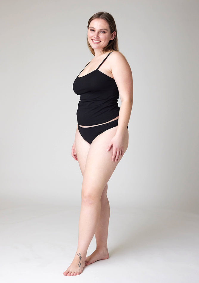 Quarter front  image of a five foot five, size 14 model wearing Ugly Pants' Black Bikini Brief period Pants in moderate  to heavy absorbency, paired with a black cami top