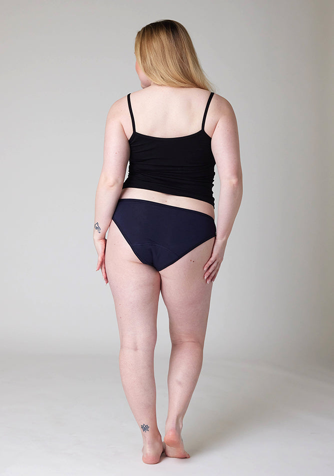 Back image of a five foot three, size 12 model wearing Ugly Pants' Navy Bikini Brief period pants in light to moderate absorbency, paired with a black cami top. 