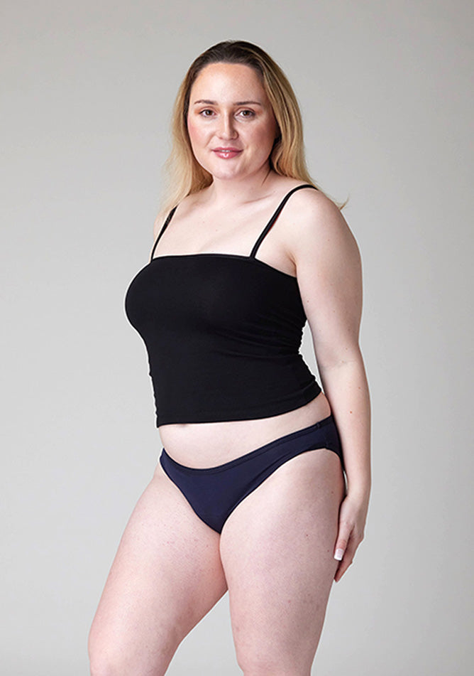 Close up quarter front image of a five foot three , size 12 model wearing Ugly Pants' Navy Bikini Brief period pants in light to moderate absorbency, paired with a black cami top. 