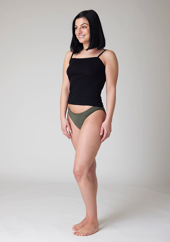 Quarter front image of a five foot six, size 8 model wearing Ugly Pants' Olive Bikini Brief period Pants in light to moderate absorbency, paired with a black cami top