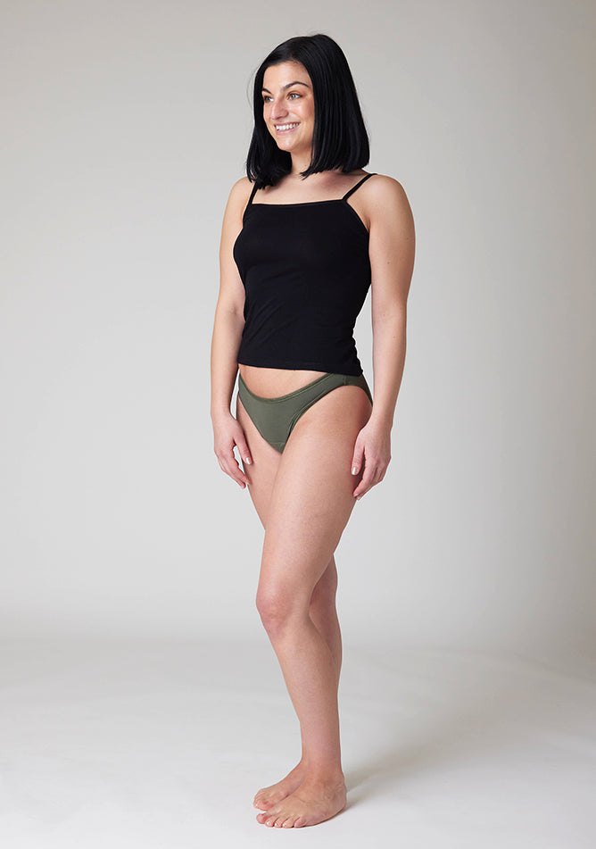 Quarter front image of a five foot six, size 8 model wearing Ugly Pants' Olive Bikini Brief period Pants in moderate  to heavy absorbency, paired with a black cami top