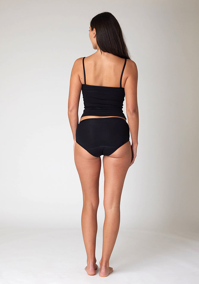 Back image of a five foot ten, size 10 model wearing Ugly Pants' Black Hipster Brief Period Pants in light to moderate  absorbency, paired with a black cami top