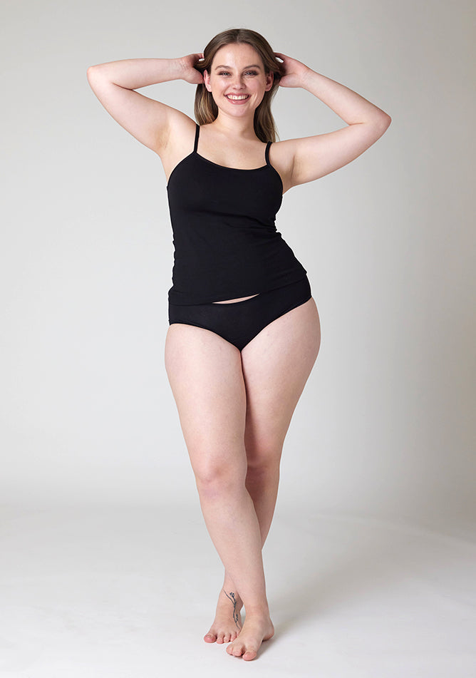 Front image of a five foot five, size 14 model wearing Ugly Pants' Black Hipster Brief Period Pants in light to moderate  absorbency, paired with a black cami top