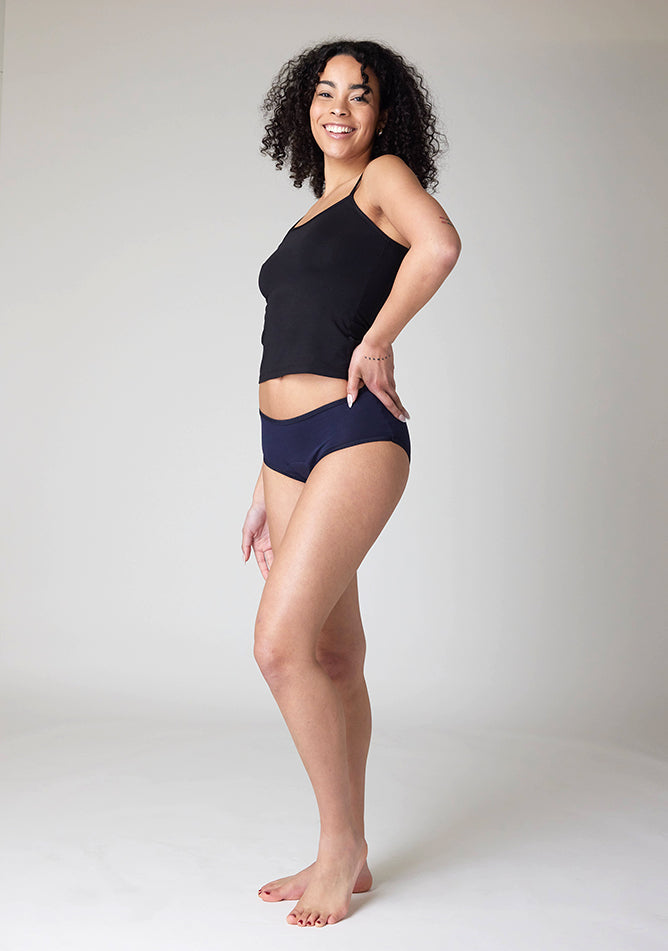 Quarter front image of a five foot ten, size 10 model wearing Ugly Pants' Navy Hipster Brief Period Pants in light to moderate  absorbency, paired with a black cami top