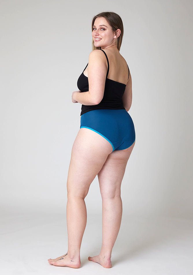 Quarter back image of a five foot five, size 14 model wearing Ugly Pants' Teal Hipster Brief Period Pants in light to moderate  absorbency, paired with a black cami top