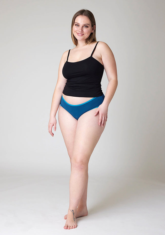 Front image of a five foot five, size 14 model wearing Ugly Pants' Teal Hipster Brief Period Pants in light to moderate  absorbency, paired with a black cami top
