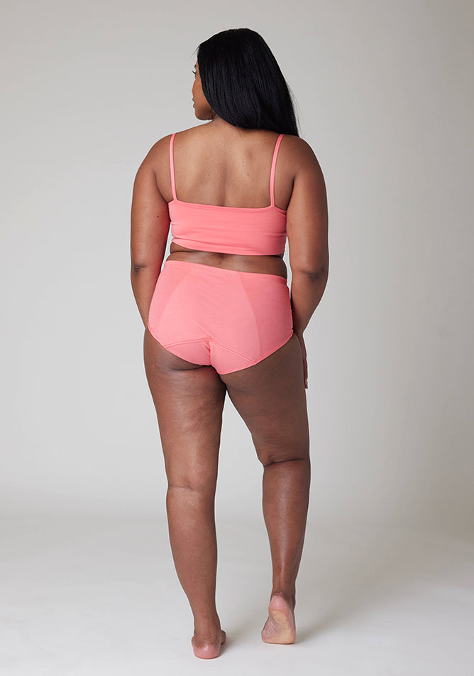 Back image of a five foot five, size 10 model wearing Ugly Pants' Coral Overnight period pants in moderate to heavy absorbency, paired with a coral crop top, this image does not feature the self tie bow, but a straight neckline. 