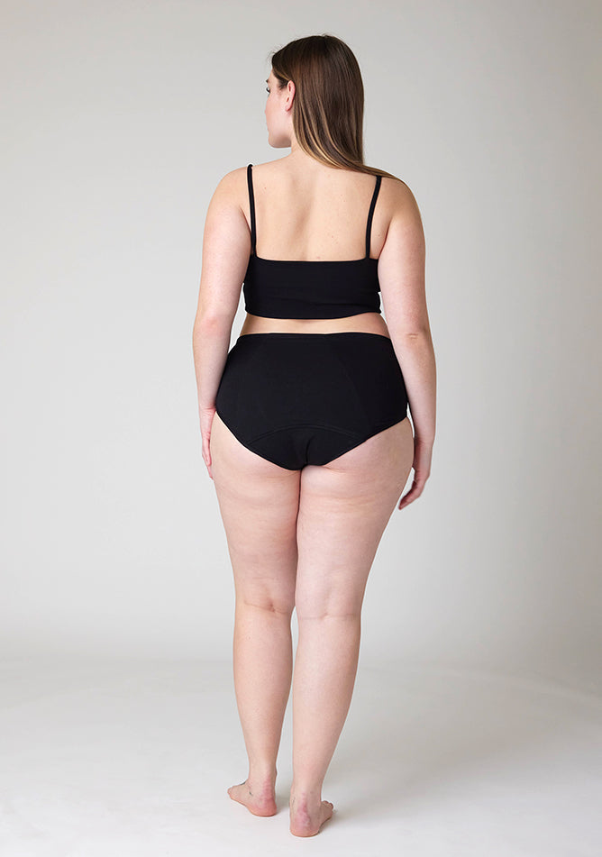 Back image of a five foot five, size 14 model wearing Ugly Pants' black Overnight period pants in moderate to heavy absorbency, paired with a black crop top, this image does not feature the self tie bow, but a straight neckline. 