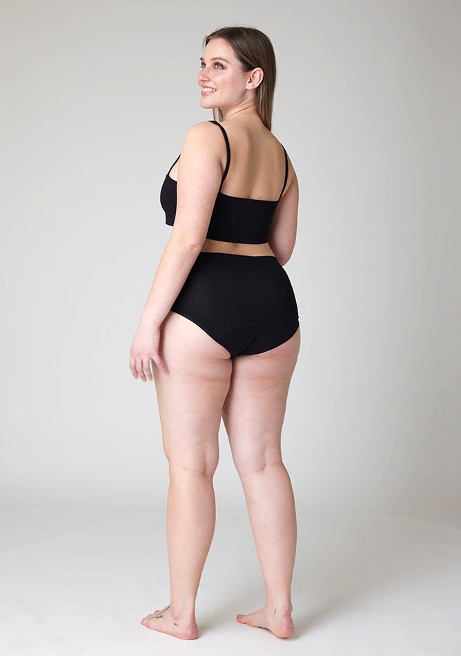 Quarter back image of a five foot five, size 14 model wearing Ugly Pants' black Overnight period pants in moderate to heavy absorbency, paired with a black crop top, this image does not feature the self tie bow, but a straight neckline. 