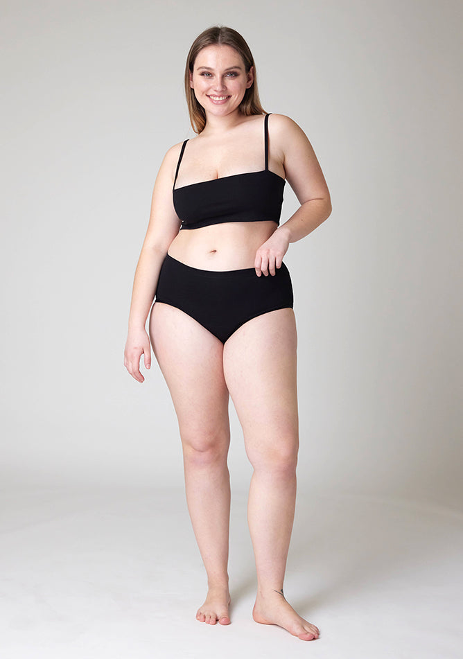 Front image of a five foot five, size 14 model wearing Ugly Pants' black Overnight period pants in moderate to heavy absorbency, paired with a black crop top, this image does not feature the self tie bow, but a straight neckline. 