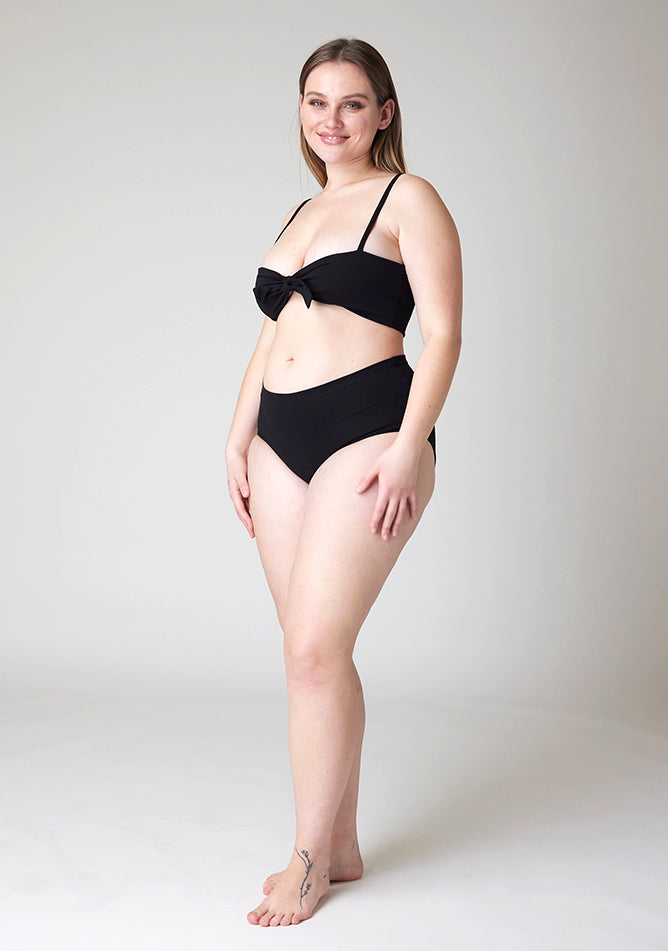 Quarter front image of a five foot five, size 14 model wearing Ugly Pants' black Overnight period pants in moderate to heavy absorbency, paired with a black crop top, this image does not feature the self tie bow.