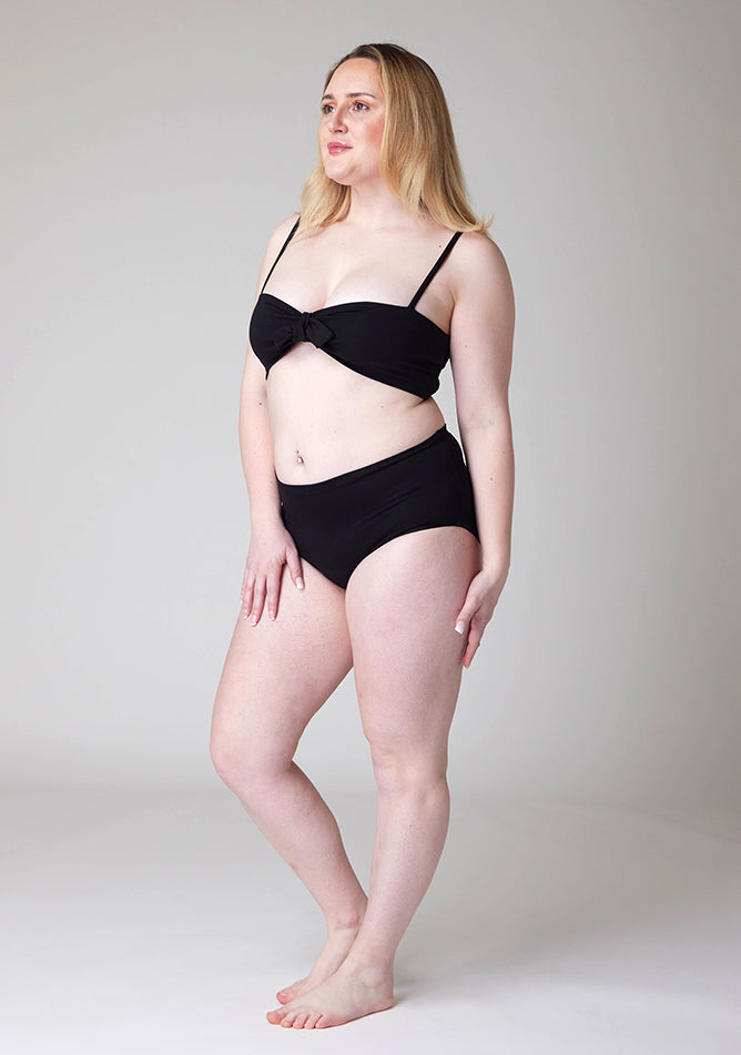 Quarter front image of a five foot three, size 12 model wearing Ugly Pants' black Overnight period pants in moderate to heavy absorbency, paired with a black crop top, this image does not feature the self tie bow.