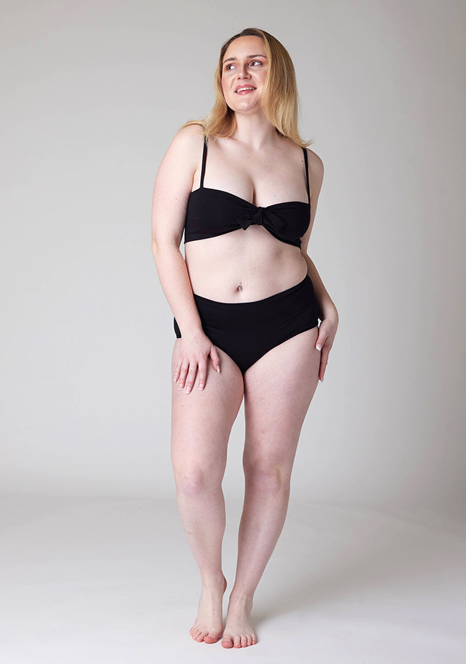 Front image of a five foot three, size 12 model wearing Ugly Pants' black Overnight period pants in moderate to heavy absorbency, paired with a black crop top, this image does not feature the self tie bow.