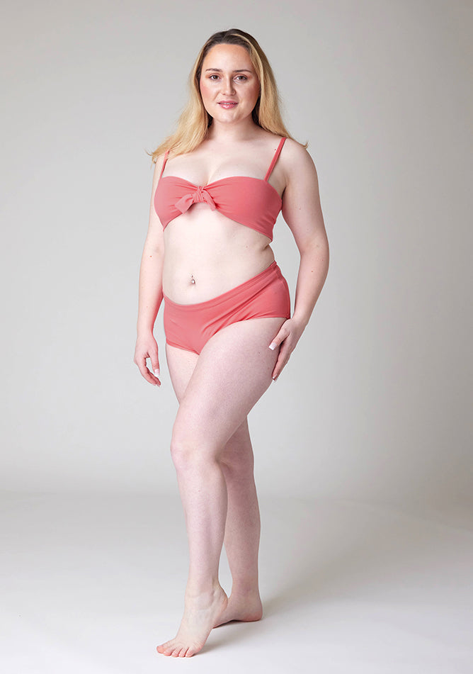 Quarter front image of a five foot three, size 12 model wearing Ugly Pants' Coral Overnight period pants in moderate to heavy absorbency, paired with a coral crop top, this image does not feature the self tie bow.