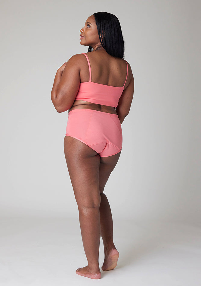 Quarter back image of a five foot five, size 10 model wearing Ugly Pants' Coral Overnight period pants in moderate to heavy absorbency, paired with a coral crop top, this image does not feature the self tie bow, but a straight neckline. 