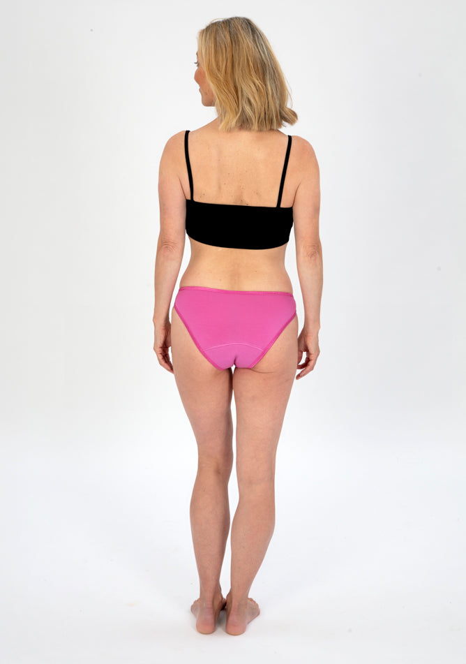 Back image of a five foot six, size 10 model wearing Ugly Pants' Pink Bikini Brief period Pants in moderate to heavy absorbency, paired with a black crop top