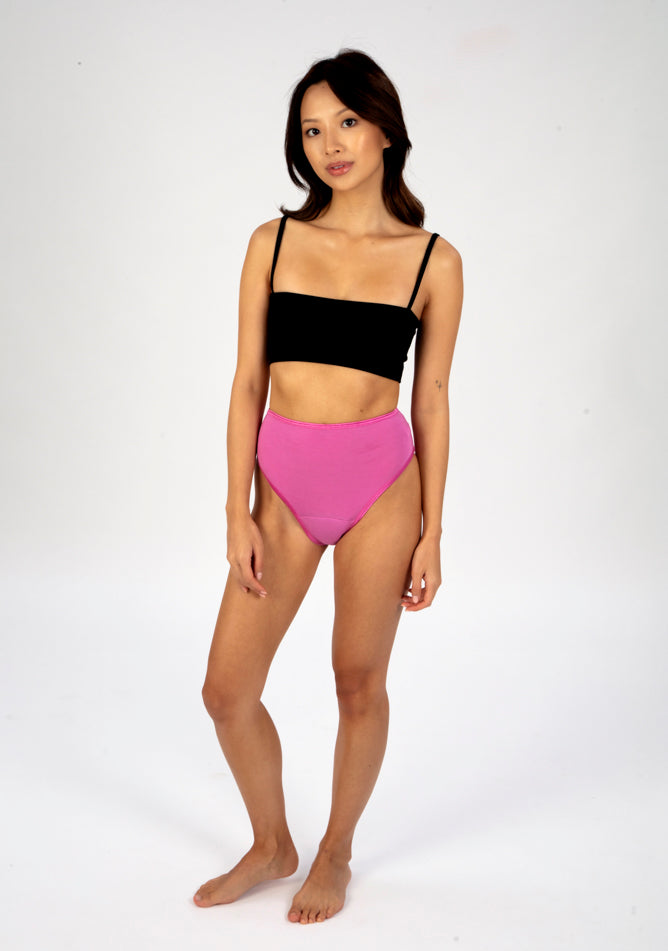 Front quarter image of a five foot three, size 6 model wearing Ugly Pants' pink high waisted period pants in light to moderate absorbency, paired with a black cami top.