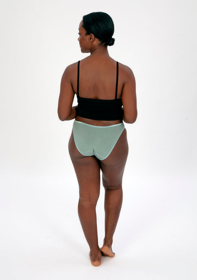 Back image of a five foot four, size 10 model wearing Ugly Pants' Sage Bikini Brief period Pants in light to moderate absorbency, paired with a black crop top