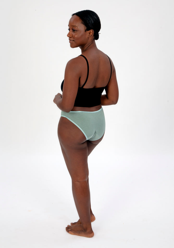 Back quarter image of a five foot four, size 10 model wearing Ugly Pants' Sage Bikini Brief period Pants in light to moderate absorbency, paired with a black crop top