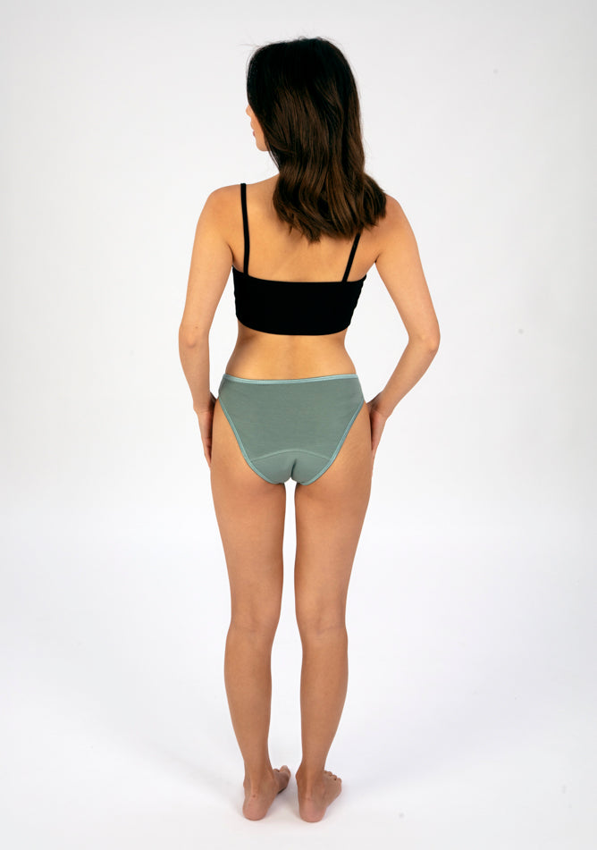 Back image of a five foot three, size 6 model wearing Ugly Pants' Sage Bikini Brief period Pants in moderate to heavy absorbency, paired with a black crop top