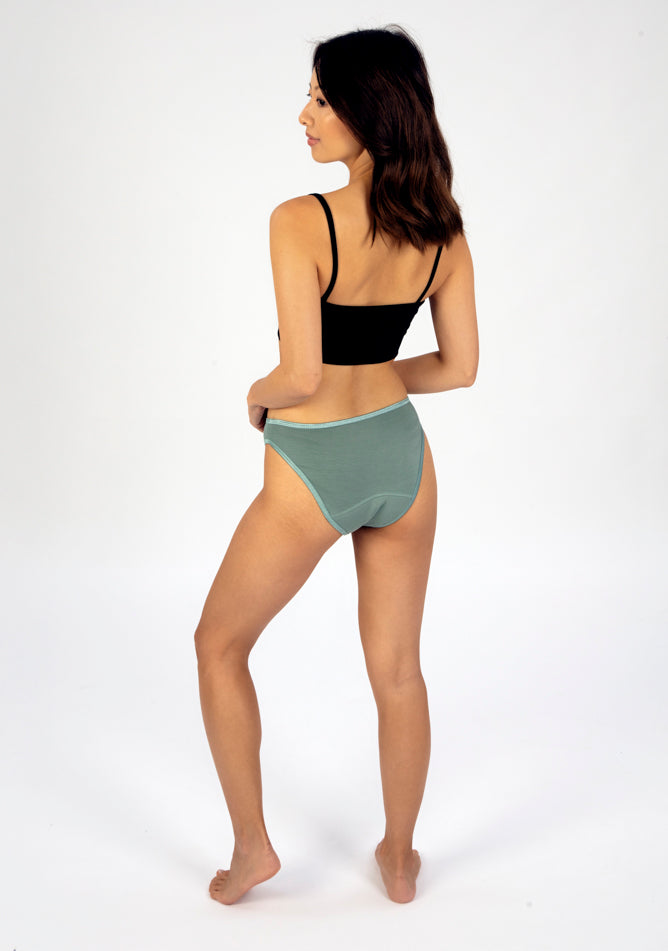 Back Quarter image of a five foot three, size 6 model wearing Ugly Pants' Sage Bikini Brief period Pants in moderate to heavy absorbency, paired with a black crop top