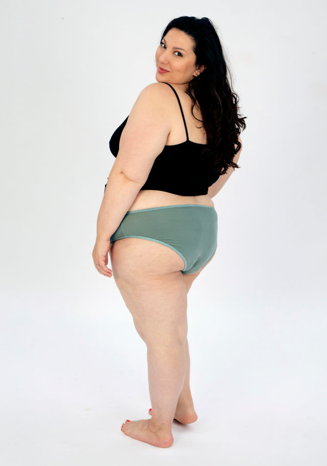 Back quarter image of a five foot seven, size 22 model wearing Ugly Pants' sage high waisted period pants in moderate to heavy absorbency, paired with a black cami top.