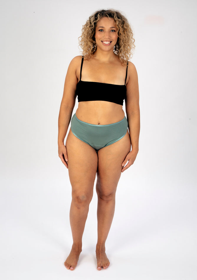 Front image of a five foot seven, size 16 model wearing Ugly Pants' sage high waisted period pants in moderate to heavy absorbency, paired with a black cami top.