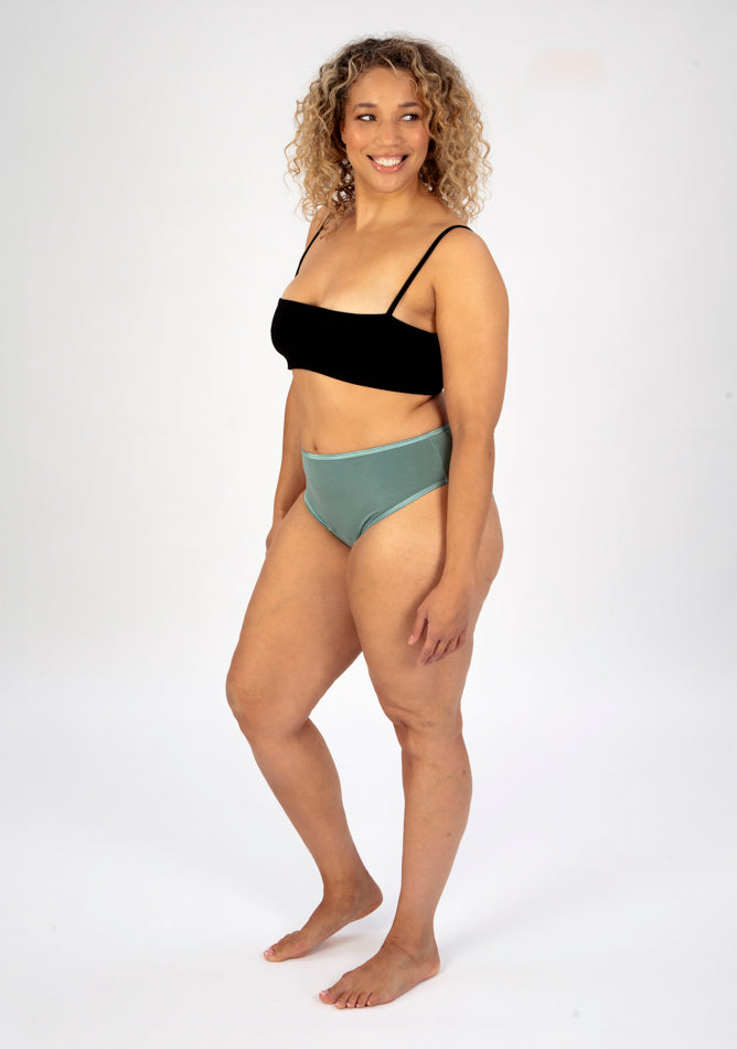 Front quarter image of a five foot seven, size 16 model wearing Ugly Pants' sage high waisted period pants in moderate to heavy absorbency, paired with a black cami top.