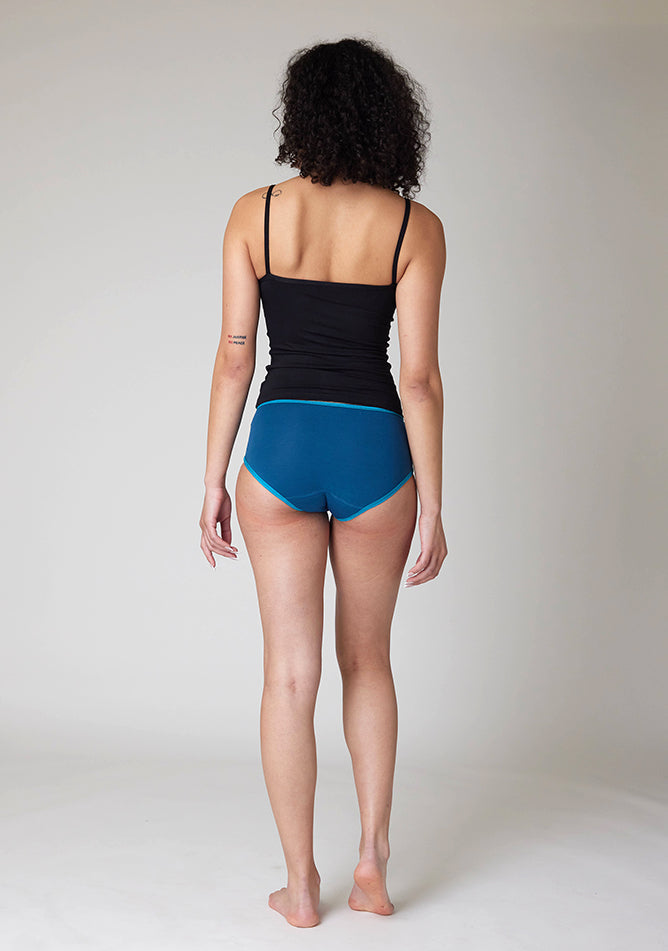 Back image of a five-foot-ten, size 10 model wearing Ugly Pants' Teal Hipster Brief Period Pants in light to moderate  absorbency, paired with a black cami top