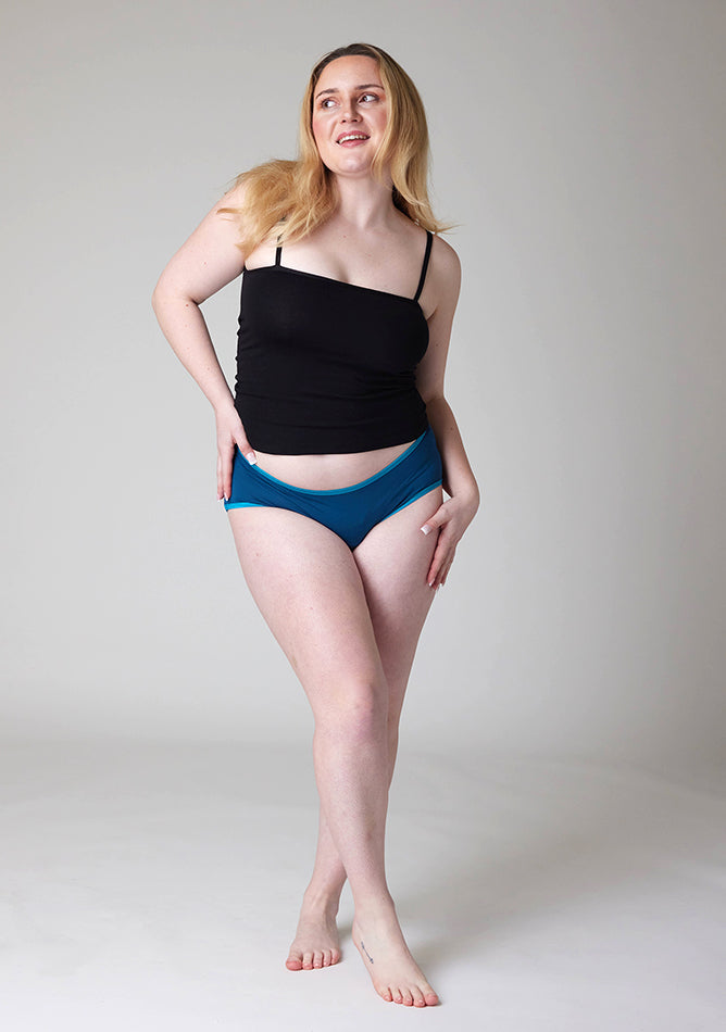Front image of a five-foot-three, size 12 model wearing Ugly Pants' Teal Hipster Brief Period Pants in light to moderate  absorbency, paired with a black cami top